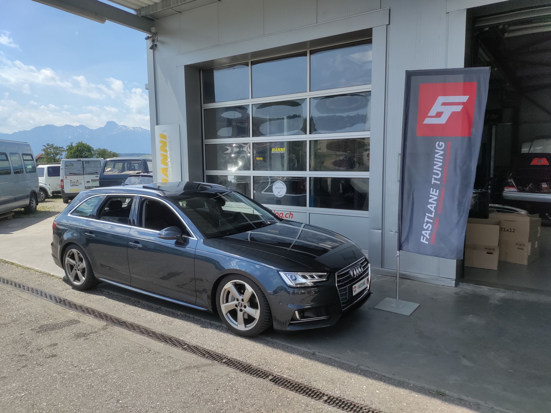 Ein dunkelgrauer Audi A6 3.0 V6 TDI Kombi steht vor der Garage neben der Fastlane Tuning Schweiz Flagge