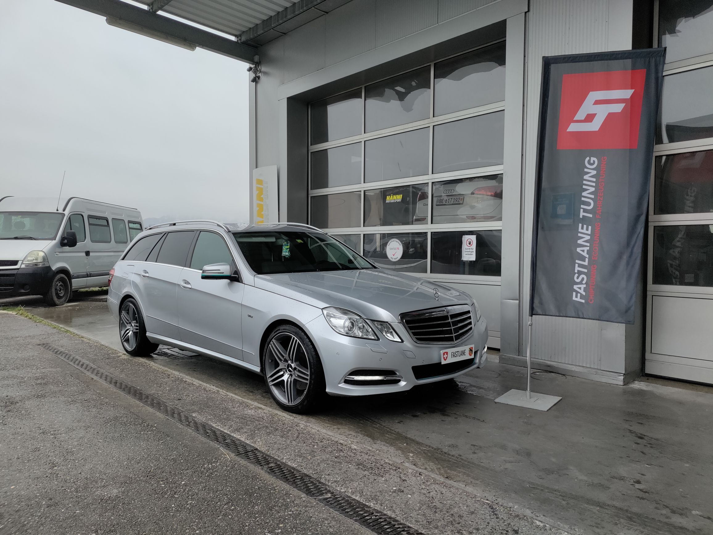 Ein Mercedes Benz E350 CDI V6 Diesel Kombi in der Farbe Silber steht vor der Garage neben der Fastlane Tuning Schweiz Flagge