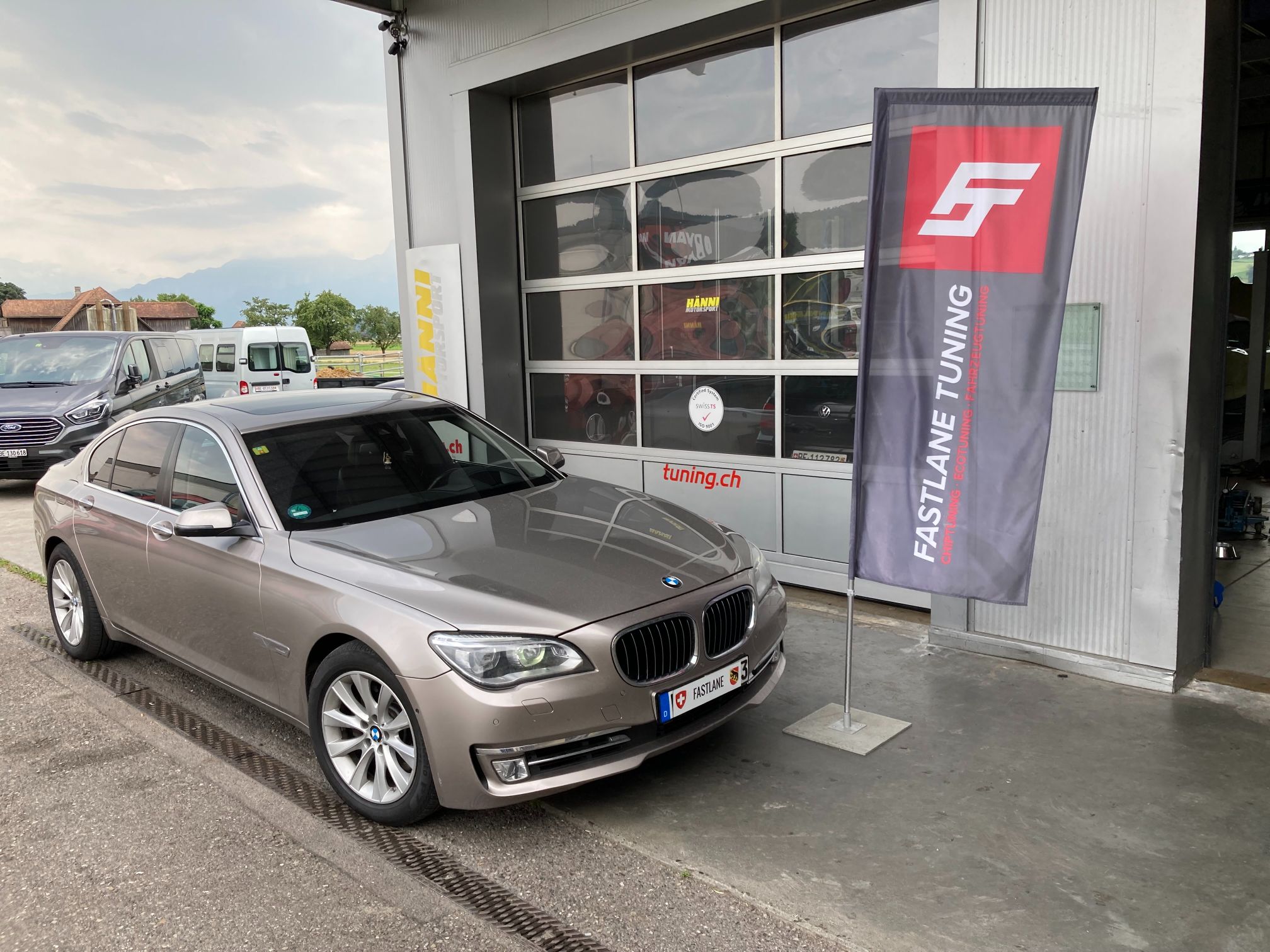 Ein hellbrauner BMW 740 Diesel steht vor der Garage neben der Fastlane Tuning Schweiz Flagge.