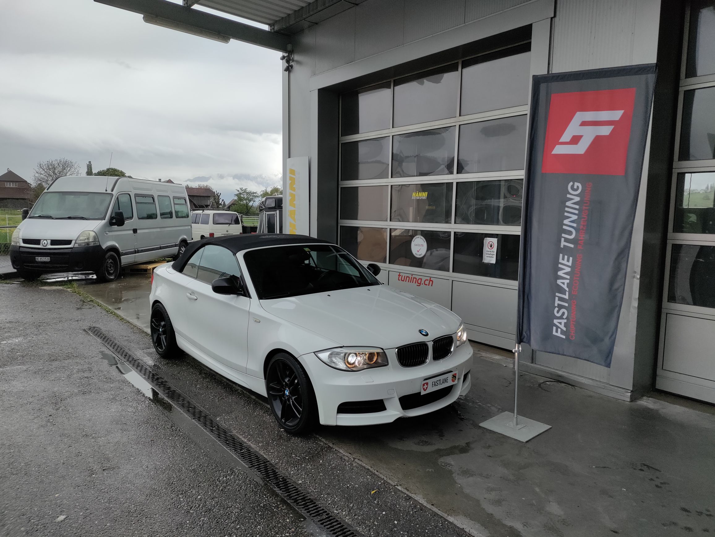 Ein weisses BMW M135i E Series Cabriolet steht vor der Garage neben der Fastlane Tuning Schweiz Flagge.
